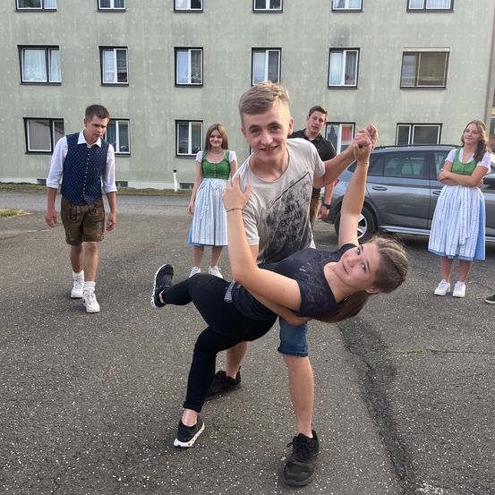 Eine Gruppe junger Leute steht vor einem Wohngebäude, einige tragen traditionelle Kleidung. Ein Paar hebt spielerisch eine Frau in die Luft.