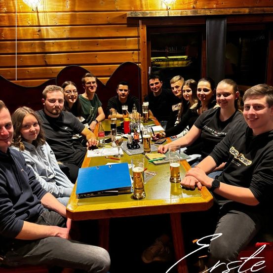 Eine Gruppe von Menschen sitzt in einem Pub um einen Tisch und posiert für ein Foto. Gläser, Bücher und Papiere liegen auf dem Tisch. Ein Wasserzeichen befindet sich unten rechts.
