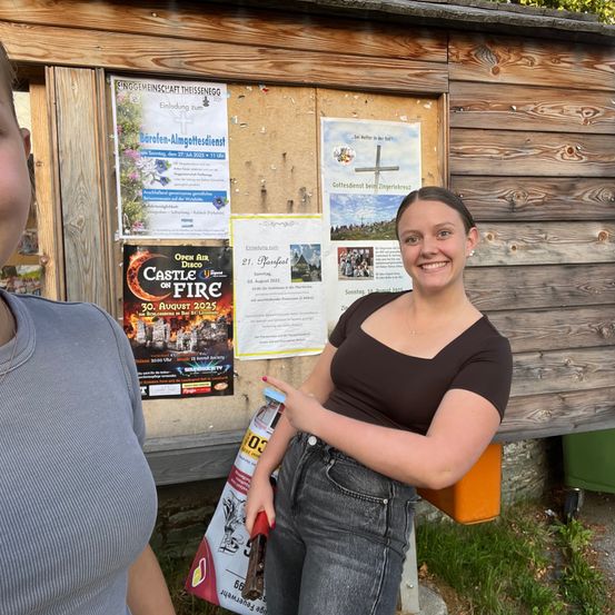 Zwei Personen stehen neben einem Holzschuppen mit Postern. Eine lächelnde Frau in Braun hält eine Flasche. Eine andere Person trägt ein graues Shirt. Poster beinhalten 'Open Air Castle on Fire' und Details zu religiösen Veranstaltungen.