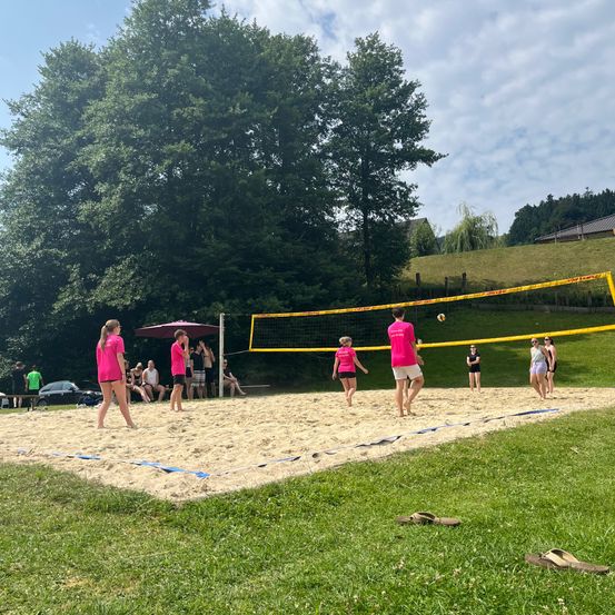 Eine Gruppe von Menschen spielt Volleyball auf einem Sandplatz. Einige Spieler tragen rosa Shirts. Zuschauer sitzen auf der Bank unter einem Schirm.