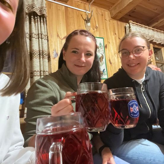 Drei Frauen sitzen und halten rote Getränke in der Hand und lächeln für die Kamera. Die Frau in der Mitte hält ein Glas mit einem Logo darauf.
