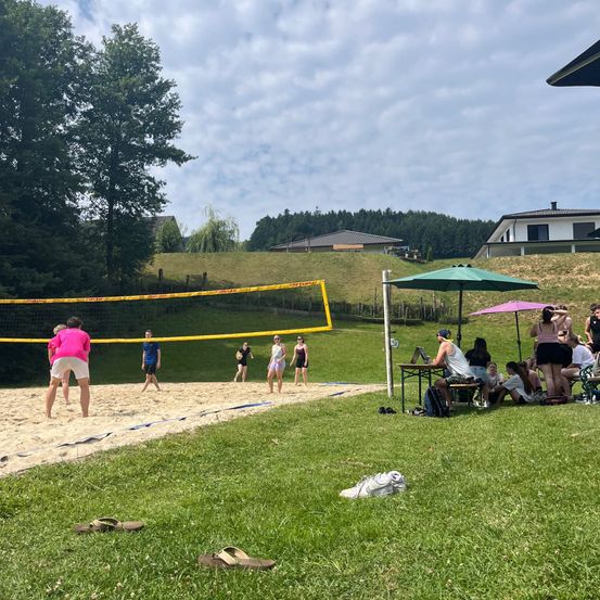 Eine Gruppe von Menschen spielt Beachvolleyball auf einem sandigen Platz, der von Gras umgeben ist. Ein gelbes Netz teilt den Platz. Zuschauer sitzen unter Schirmen auf der Rasenfläche.