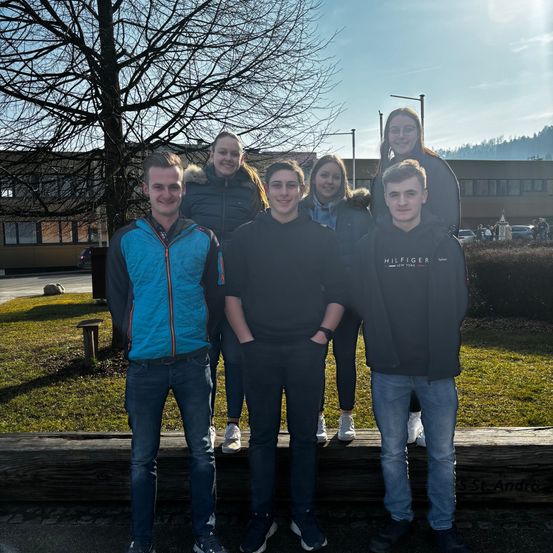 Eine Gruppe junger Leute steht auf einer Bank vor einem Gebäude. Dahinter befindet sich ein Baum und ein Rasenbereich. Das Wetter ist sonnig.