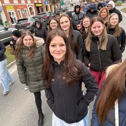 Eine Gruppe von Frauen in Winterjacken steht auf einer Straße, eine lächelnde Frau in der Mitte. Autos sind am Straßenrand geparkt. Gebäude und Bäume im Hintergrund.