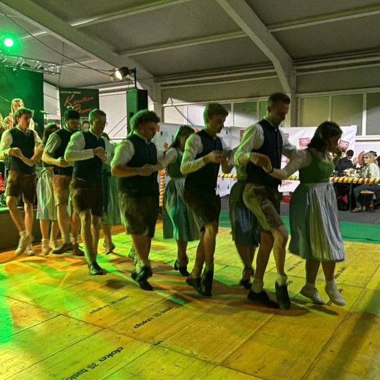 Eine Gruppe von Menschen in traditioneller Kleidung tanzt auf einer Bühne. Ein grünes Licht und Lautsprecher sind sichtbar. Das Publikum sitzt hinter einer Barriere.