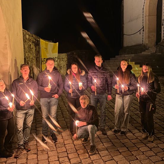 Eine Gruppe von Menschen mit brennenden Kerzen steht nachts auf einer Kopfsteinpflasterstraße. Sie sind warm gekleidet und von ihren Kerzen beleuchtet.