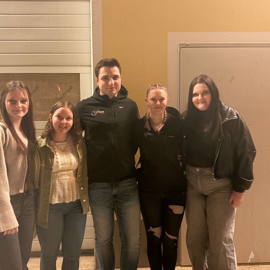 Fünf Menschen stehen lächelnd vor einem Gebäude. Der Mann in der Mitte trägt eine schwarze Jacke mit Logo. Die Frauen tragen verschieden farbige Jacken.