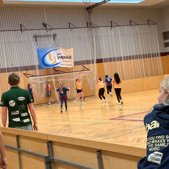 Indoor-Spielfeld mit aktiven Spielern. Ein Banner mit Jugend steht über dem Tor. Zuschauer beobachten von der Seite.