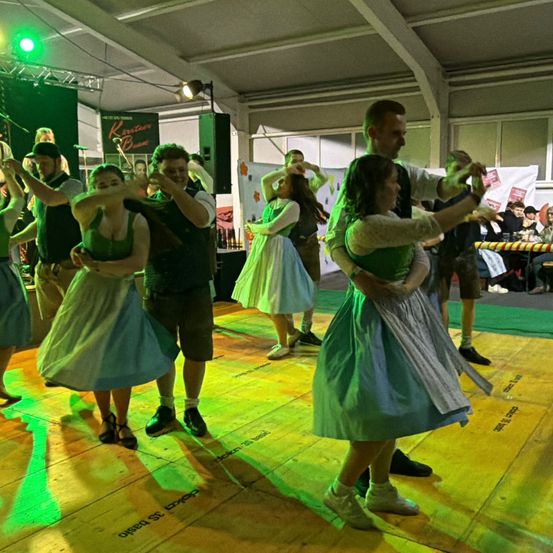 Eine Gruppe von Menschen tanzt auf einem Holzboden unter grünem Licht. Ein Mann in einem schwarzen Hemd hält eine Frau in einem grünen Kleid.