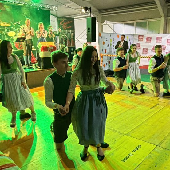 Ein Paar tanzt auf einer Bühne, die von grünen Lichtern beleuchtet wird, während eine Band hinter ihnen spielt. Andere schauen zu und tanzen in traditioneller Kleidung.