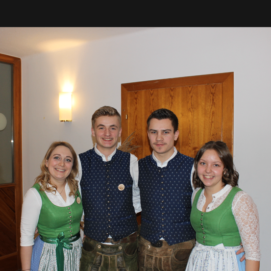 Vier Personen, zwei Frauen und zwei Männer, lächeln für die Kamera. Sie tragen traditionelle deutsche Kleidung. Hinter ihnen befindet sich eine weiße Wand mit einer Holztür.