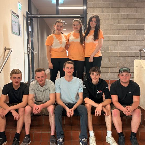 Eine Gruppe von sieben jungen Erwachsenen, drei Männer und vier Frauen, lächeln für ein Foto auf einer Treppe. Sie tragen passende orangefarbene und weiße Shirts.