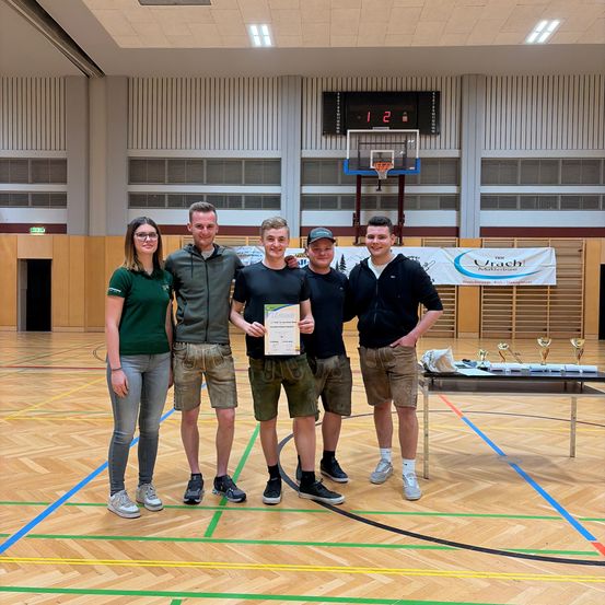 Fünf Personen stehen in einer Turnhalle mit einem Basketballfeld im Hintergrund. Drei Männer und zwei Frauen lächeln. Ein Mann hält eine Urkunde.