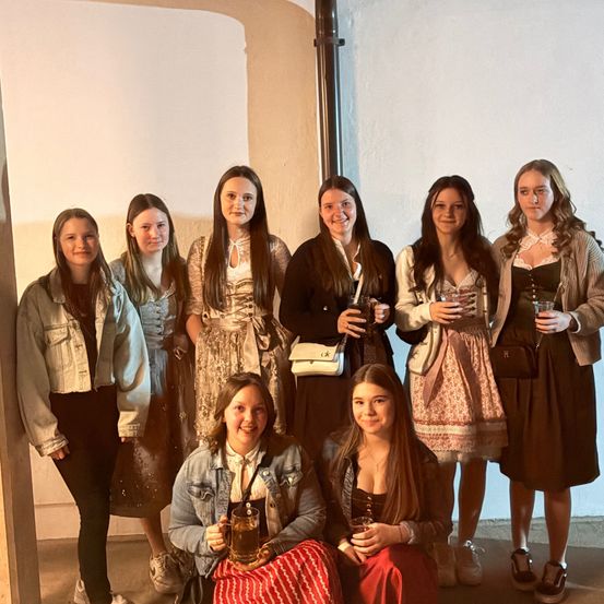 Eine Gruppe junger Frauen in traditioneller und lässiger Kleidung posiert für ein Foto und hält Getränke in der Hand. Sie lächeln und stehen eng zusammen vor einer schlichten weißen Wand.