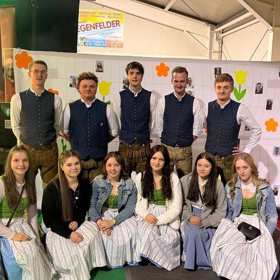 Eine Gruppe von Menschen in traditioneller bayerischer Kleidung posiert für ein Foto vor einem Hintergrund mit einem Plakat und verschiedenen Bildern.