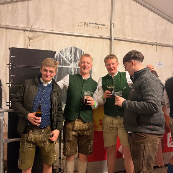 Vier junge Männer in traditioneller bayerischer Kleidung, stehen zusammen mit Getränken in der Hand, lächeln und schauen in die Kamera in einer festlichen Umgebung.