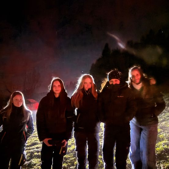 Fünf Personen, eingehüllt in Jacken, stehen zusammen in einem schwach beleuchteten Bereich. Sie scheinen für ein Foto zu lächeln, mit einem dunklen, rauchigen Hintergrund hinter ihnen.