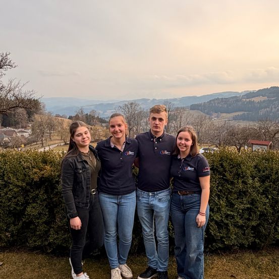 Vier Personen, zwei Männer und zwei Frauen, posieren für ein Foto vor einer Hecke, mit Bergen im Hintergrund.