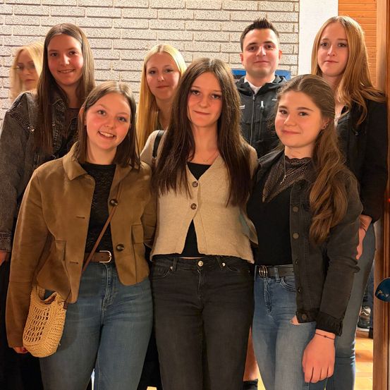 Eine Gruppe junger Menschen posiert für ein Foto, lächelnd. Eine Person hält eine Tasche, eine andere hat eine Halskette. Eine Ziegelmauer ist im Hintergrund.