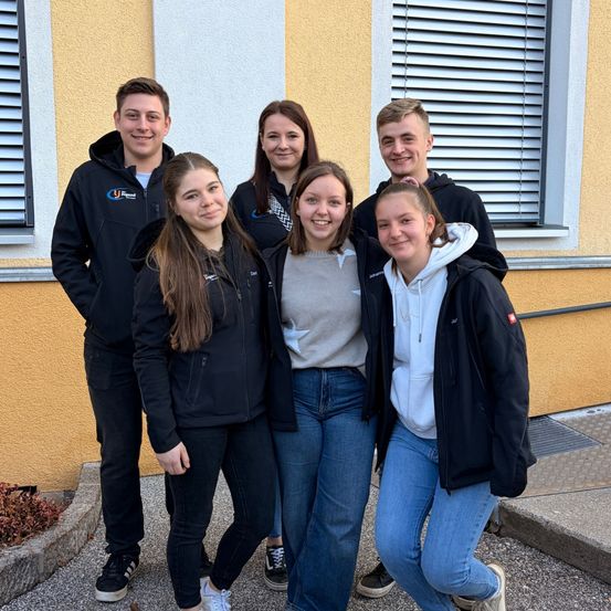 Fünf Personen stehen lächelnd vor einem gelben Gebäude mit Fenstern. Sie tragen Jacken und Turnschuhe. Ein Junge vorne trägt eine Kapuzenjacke.