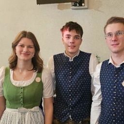 Drei Personen stehen zusammen, zwei Männer und eine Frau. Die Frau trägt ein grünes Kleid mit Knöpfen und beide Männer tragen Westen. Der Mann auf der rechten Seite trägt eine Brille. Sie stehen vor einer weißen Wand.