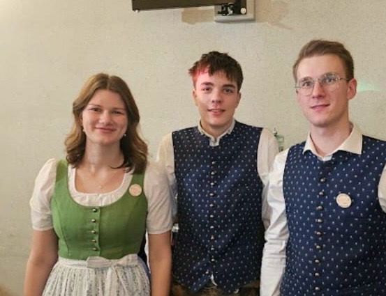 Drei Personen stehen zusammen, zwei Männer und eine Frau. Die Frau trägt ein grünes Kleid mit Knöpfen und beide Männer tragen Westen. Der Mann auf der rechten Seite trägt eine Brille. Sie stehen vor einer weißen Wand.