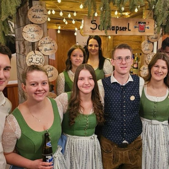 Eine Gruppe von Menschen in traditioneller bayerischer Kleidung posiert für ein Foto, mit Schildern auf Deutsch und einer festlichen Atmosphäre.