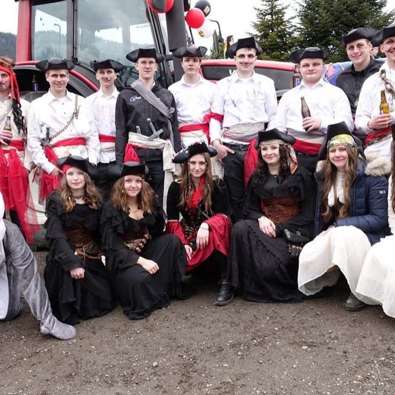 Eine Gruppe von Menschen in Kostümen posiert für ein Foto mit einem Traktor im Hintergrund. Einige halten Flaschen und tragen Hüte.