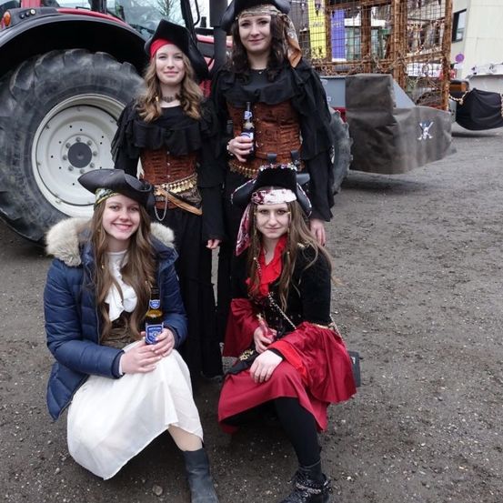 Vier Frauen in Piratenkostümen posieren lächelnd vor einem Traktor. Eine hält eine Bierflasche.