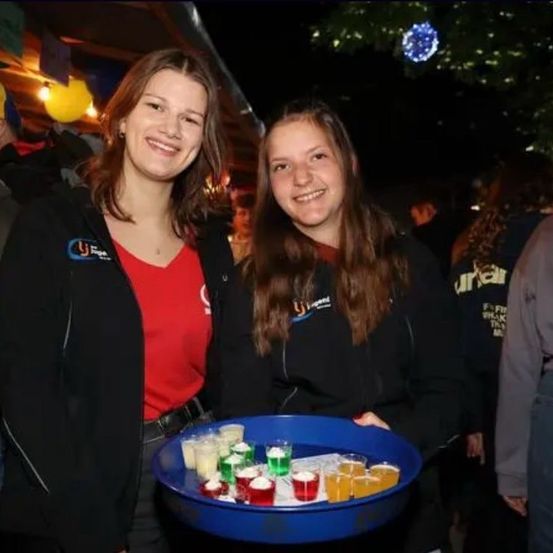 Zwei Frauen lächeln und posieren für ein Foto. Eine hält einen Tablett mit Getränken, während die andere hinter ihr steht. Im Hintergrund sind andere Menschen und festliche Dekorationen zu sehen.