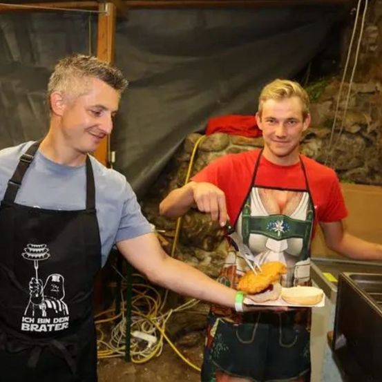 Zwei Männer in Schürzen stehen in einem Raum, einer hält einen Teller mit Essen und der andere einen Messer. Eine Frau mit einer Star Wars Schürze lächelt hinter ihnen.