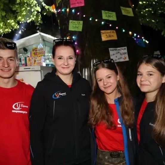 Vier junge Leute stehen zusammen in einer festlichen, outdoor-Umgebung und lächeln in die Kamera. Hinter ihnen sind Bäume, die mit bunten Lichtern und Schildern geschmückt sind, eines davon steht 'Happy Hour'.