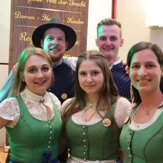 Vier Frauen in grünen Kleidern mit weißen Spitzenbesätzen posieren für ein Foto. Hinter ihnen steht ein Holzschild mit dem Text 'Damen-He' und 'Reg' darauf. Die beiden Männer im Hintergrund lächeln und posieren mit ihren Armen um die Frauen.