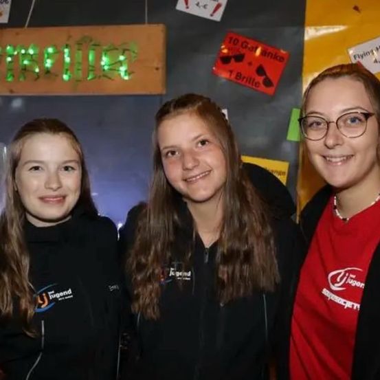 Drei junge Frauen stehen zusammen und lächeln. Sie tragen schwarze Jacken mit Logos. Hinter ihnen zeigt eine Wand Schilder und ein Holzschild mit beleuchteten Buchstaben.
