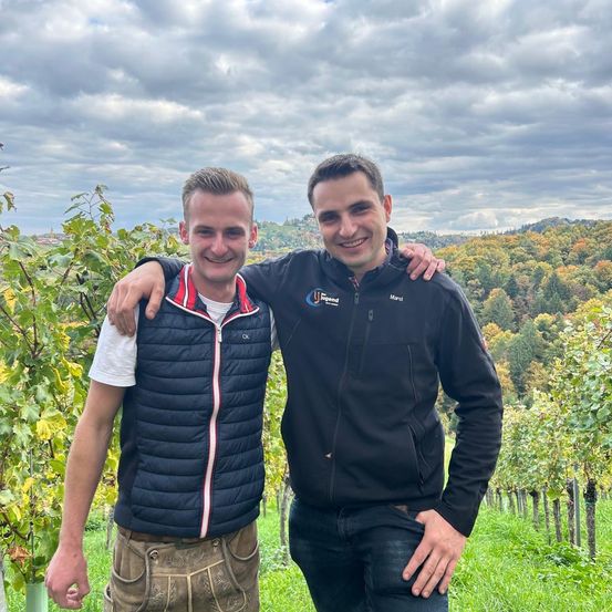 Zwei Männer stehen in einem Weinberg und lächeln für die Kamera. Einer trägt eine blaue Weste, der andere eine schwarze Jacke mit 'Legend' darauf. Bäume und Berge bilden den Hintergrund.