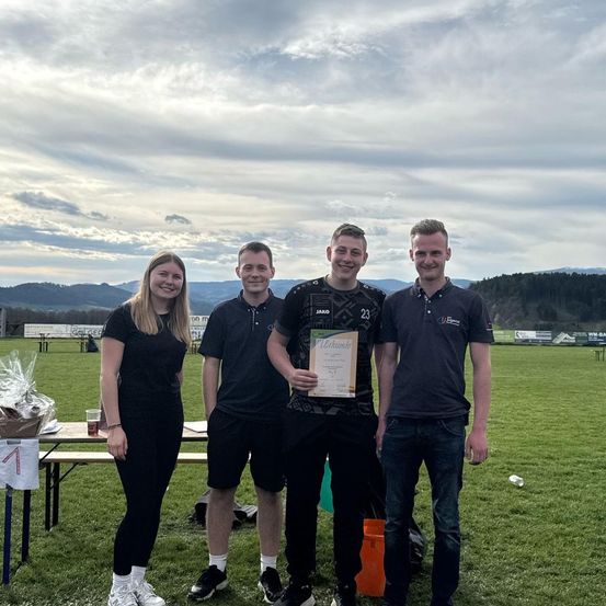 Vier Menschen stehen auf einem Rasenfeld mit Bergen im Hintergrund und lächeln für ein Foto. Der Mann in der Mitte hält ein Zertifikat.