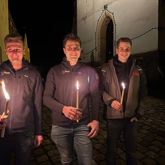 Drei Männer stehen zusammen, halten brennende Kerzen, lächeln und posieren vor einem alten Gebäude bei Nacht.