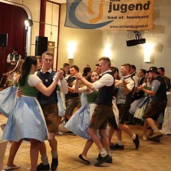 Eine Gruppe von Menschen tanzt in einem Saal, gekleidet in traditioneller Kleidung. Ein Banner mit 'Jugend' hängt oben. Lautsprecher und Lichter sind sichtbar.