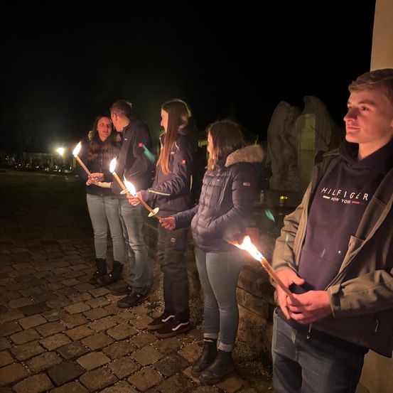 Fünf Menschen halten brennende Kerzen in einem schwach beleuchteten Außenbereich bei Nacht. Sie stehen auf einem Kopfsteinpflasterweg.