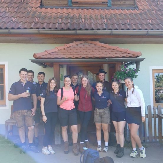 Eine Gruppe von Menschen steht vor einem Haus mit einem braunen Dach. Einige tragen Freizeitkleidung, während andere Sportbekleidung tragen. Sie scheinen für ein Gruppenfoto zu lächeln.