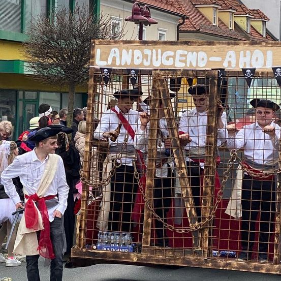 Eine Gruppe von Menschen, die als Piraten verkleidet sind, ist bei einer Parade. Sie befinden sich in einem Metallkäfig mit einem Schild, auf dem Landjugend-Aho steht.