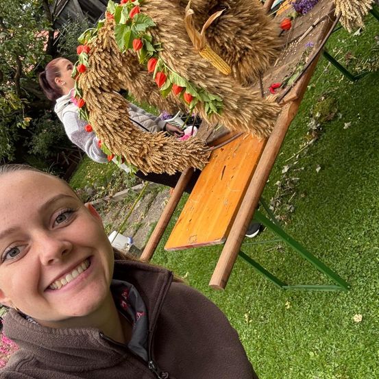 Ein lächelndes Mädchen in einer braunen Jacke posiert vor einem Holztisch mit einem Weizenkranz und Blumen. Eine andere Person ordnet im Hintergrund Blumen auf dem Tisch an.
