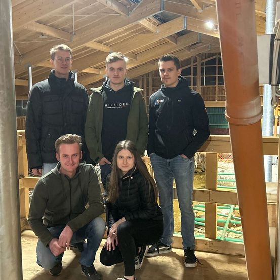 Fünf Personen posieren für ein Foto in einer Scheune mit Holzzäunen. Die Frau und der Mann hocken, während andere hinter ihnen stehen. Durch den Zaun ist eine Kuh zu sehen.