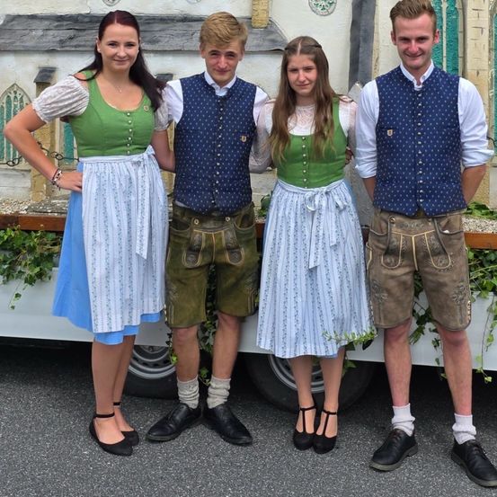 Vier Personen, zwei Männer und zwei Frauen, in traditioneller Kleidung, stehen zusammen für ein Foto. Sie stehen vor einem Gebäude mit einer dekorativen Wand und Grünpflanzen.