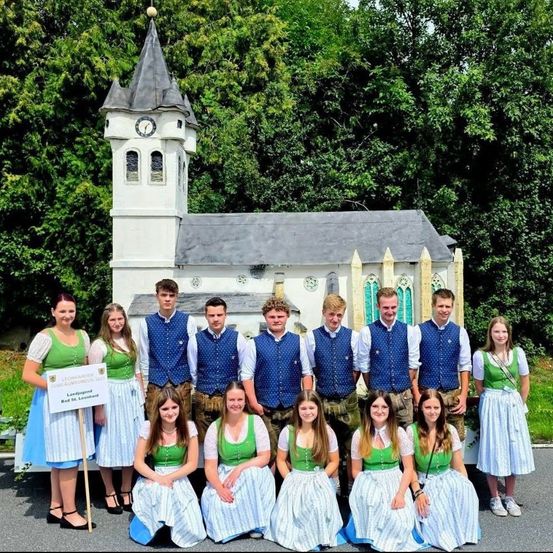Eine Gruppe junger Leute in traditioneller Kleidung posiert für ein Foto vor einer kleinen Kirche. Die Frau hält ein Schild, während Bäume und Grünpflanzen im Hintergrund sind.