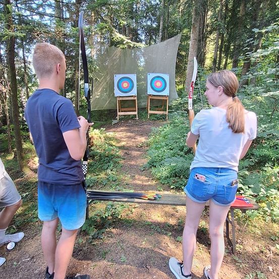 Zwei Personen üben im Wald Bogenschießen mit zwei Zielscheiben. Der Mann ist kurz vor dem Schuss, während die Frau zusieht. In der Nähe steht ein Tisch mit ihrer Ausrüstung.