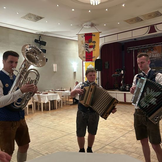Drei Männer spielen Musikinstrumente in einem Saal. Einer spielt Tuba, die anderen Akkordeons. Ein Banner hängt über ihnen, und Tische mit Stühlen sind im Hintergrund.