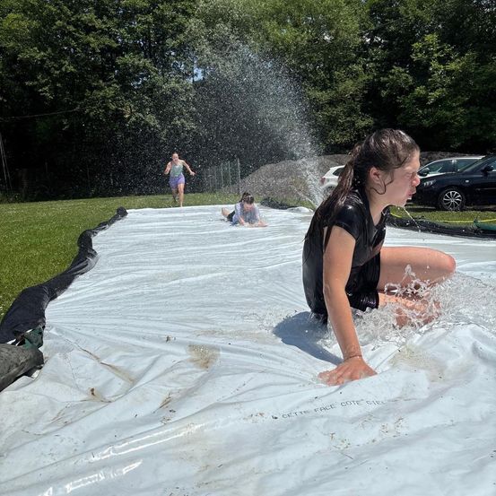 Drei junge Mädchen spielen auf einer Wasserrutsche im Garten, eine rutscht hinunter, während die anderen zusehen.