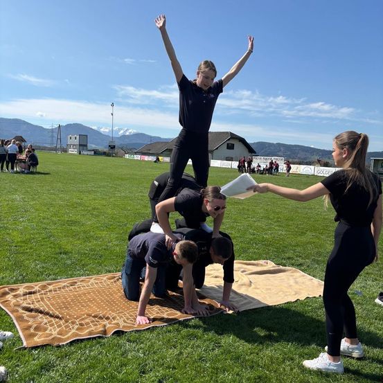 Drei Menschen bilden auf einem grünen Feld eine Pyramide, wobei eine Frau oben steht. Eine Frau hält ein Papier über der Pyramide. Das Feld ist von Bergen umgeben.