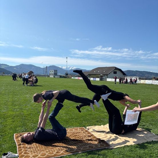 Zwei Personen führen Akrobatik auf einem Teppich in einem Grasfeld vor, während andere im Hintergrund zusehen. Berge sind in der Ferne sichtbar.